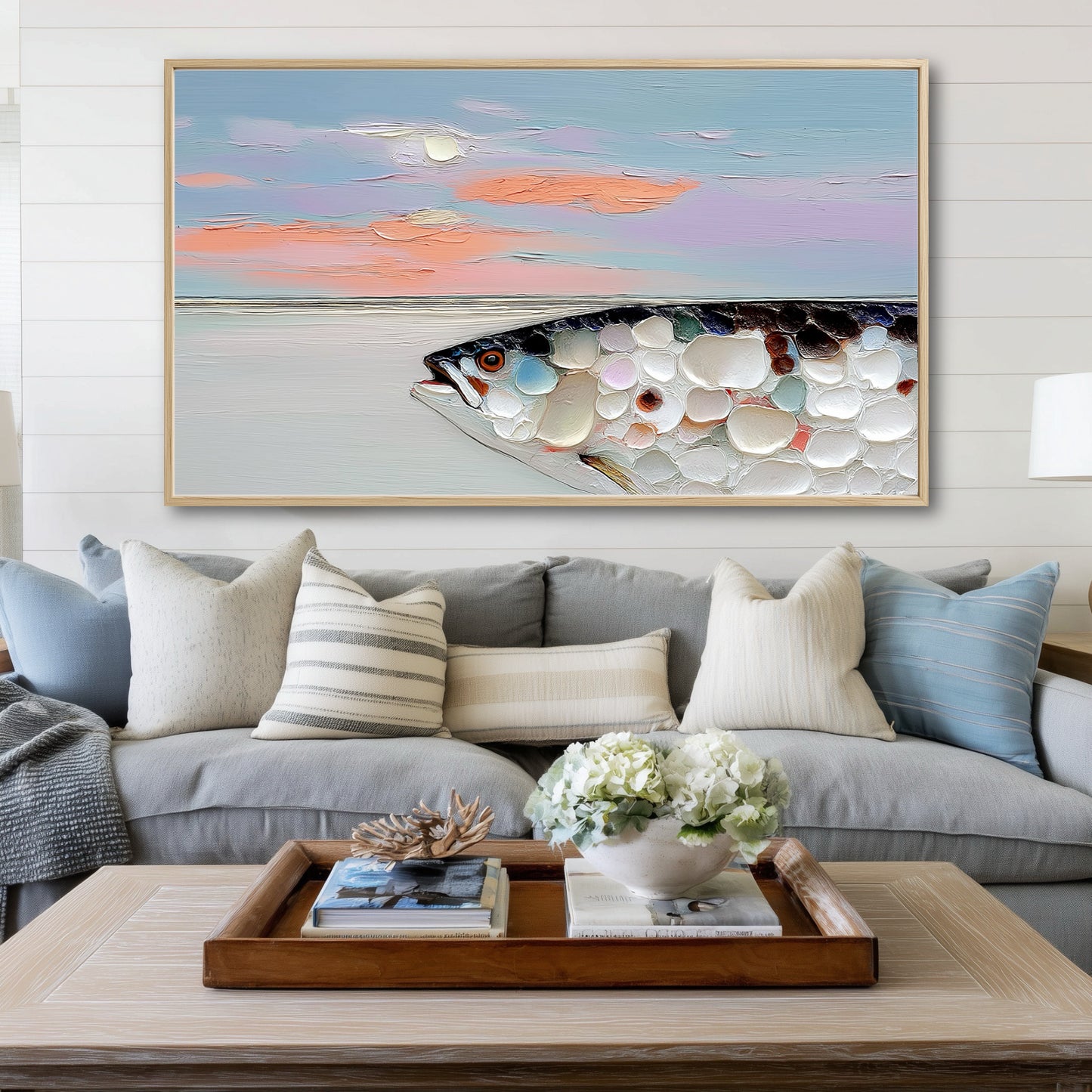 A large impasto fish painting hangs on a white shiplap wall above a gray sofa with blue and white pillows.