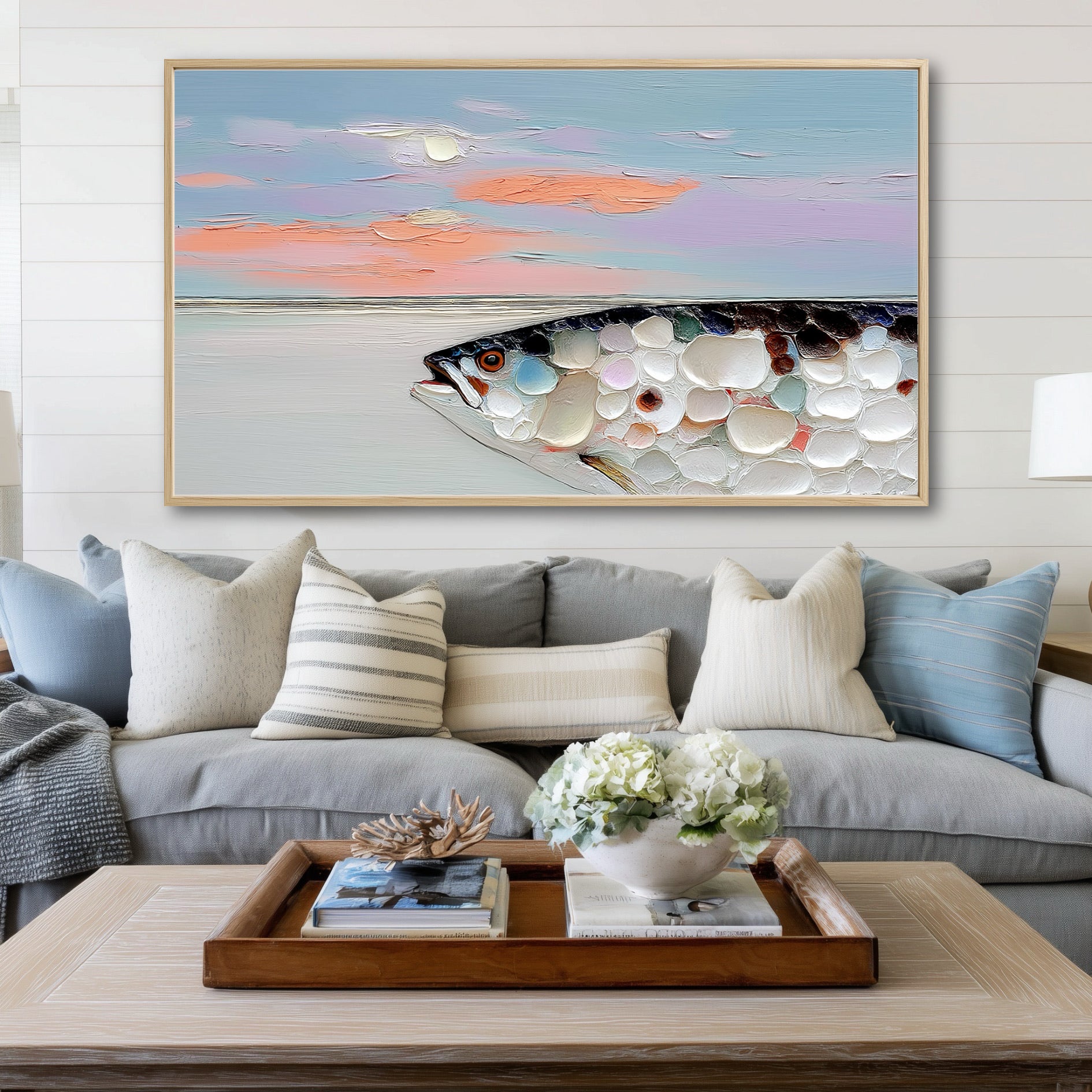 A large impasto fish painting hangs on a white shiplap wall above a gray sofa with blue and white pillows.