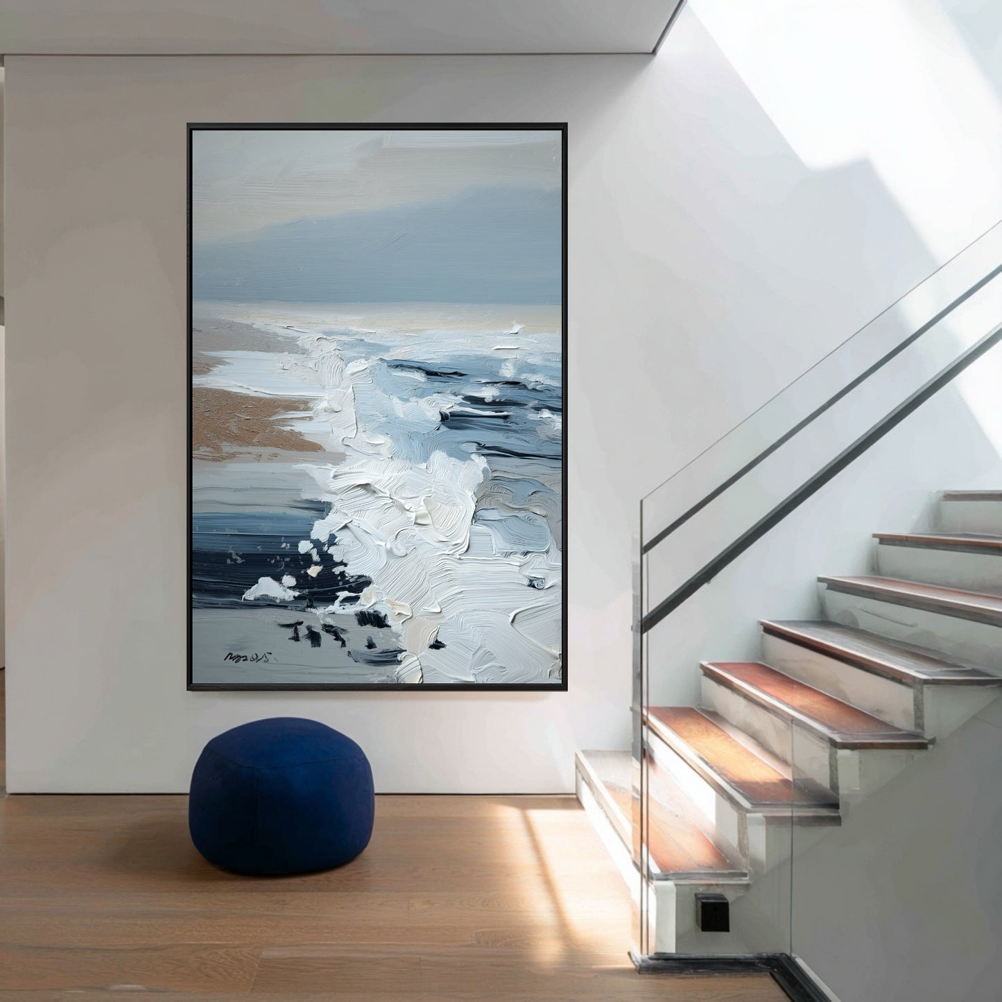 A large vertical coastal painting hangs in a modern hall, its deep blue tones perfectly matching the pouf by the staircase.