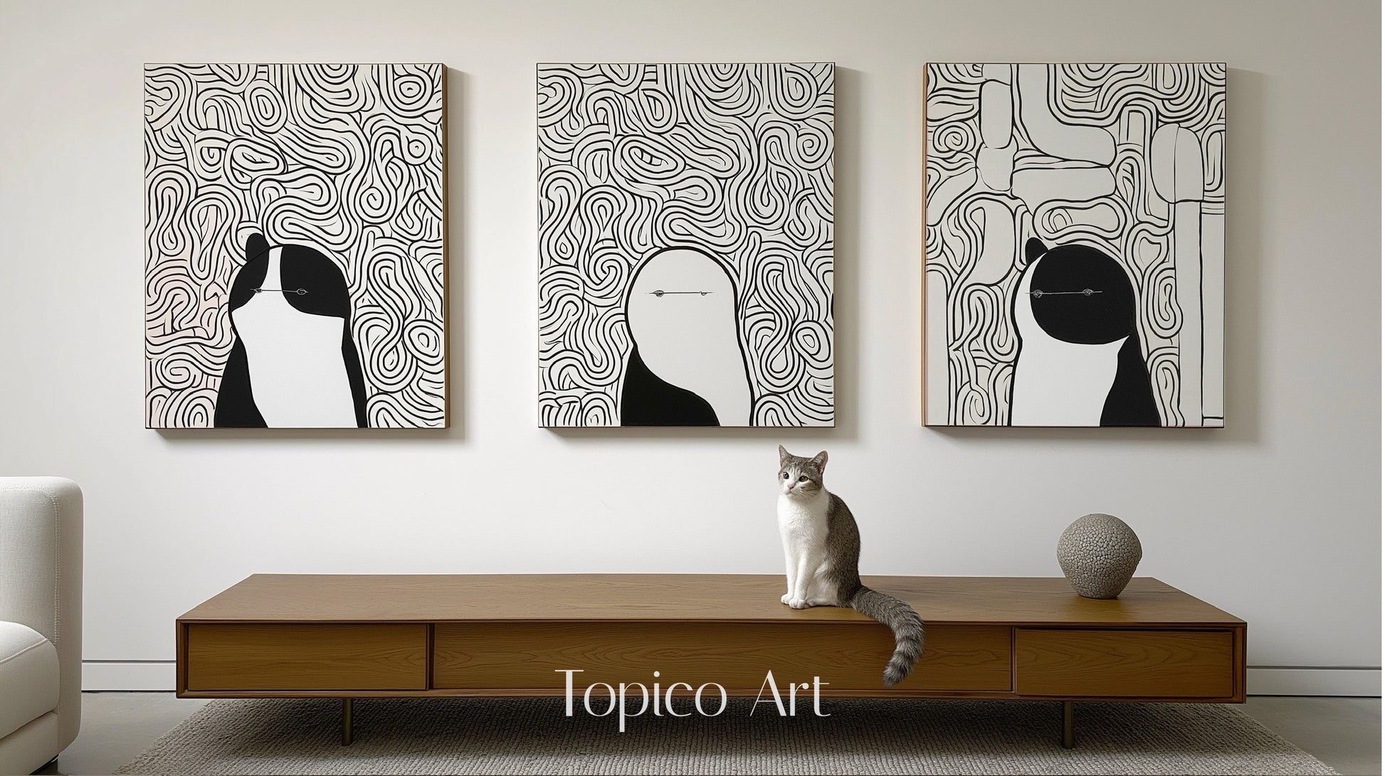 Three modern black and white canvases featuring abstract figures/animals, centered over a wood console table with a gray and white cat.