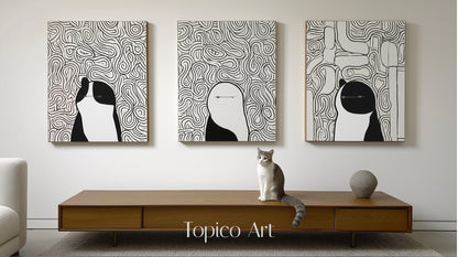 Three modern black and white canvases featuring abstract figures/animals, centered over a wood console table with a gray and white cat.