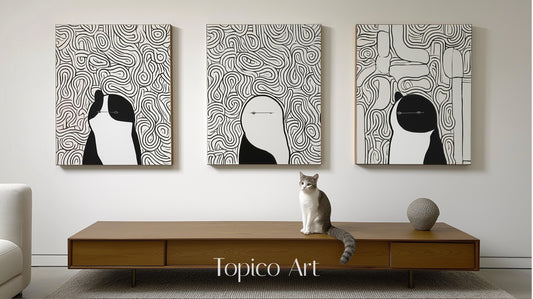 Three modern black and white canvases featuring abstract figures/animals, centered over a wood console table with a gray and white cat.